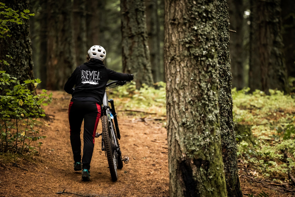 Rider walking a mountain bike through the woods in black Revel Rider Shuttle Hoodie
